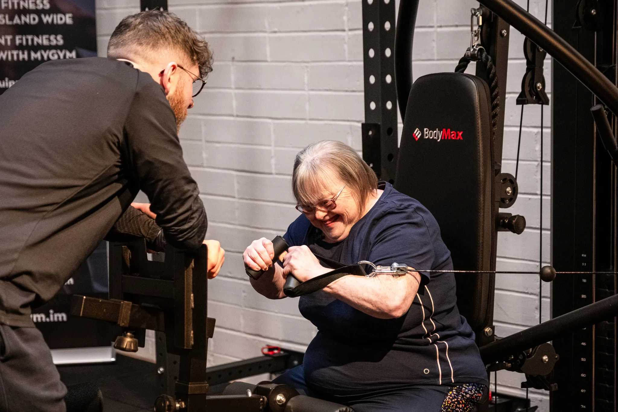 A woman with Down's syndrome smiling while using a BodyMax cable resistance machine assisted by a personal trainer in a gym, with a MyGym Isle of Man banner visible in the background